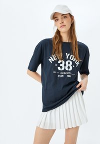 Camiseta de algodón azul marino con texto gráfico blanco "NEW YORK 38" y fecha. Combinada con una falda plisada blanca, mostrando un atuendo casual.