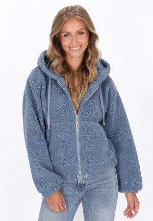 Jongeman, glimlachend, gekleed in een blauwe ritssluiting sherpa hoodie met capuchon en lichtblauwe spijkerbroek, staande tegen een witte achtergrond.