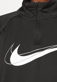 Musta urheilullinen puolivetoketjullinen pusero, jossa on korkea kaulus; siinä on suuri valkoinen Nike-swoosh-logo rinnassa; teksturoitu kangas.