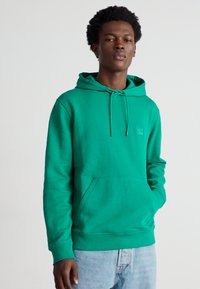 Green cotton hoodie with a front pocket, drawstring hood, and ribbed cuffs. Smooth texture with a subtly branded logo on the chest.