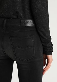 Jeans de mezclilla negra de cintura alta, con un parche de cuero en el logo y costuras de bolsillo trasero distintivas en un patrón en zigzag.
