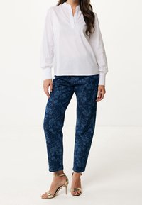 Femme portant une blouse blanche à manches longues, un jean à motifs floraux bleu marine, et des sandales à talons hauts dorées, se tenant devant un fond blanc.