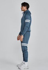 Hooded tracksuit in muted blue fabric featuring white stripes on sleeves and pants, with a fitted silhouette and kangaroo pocket.