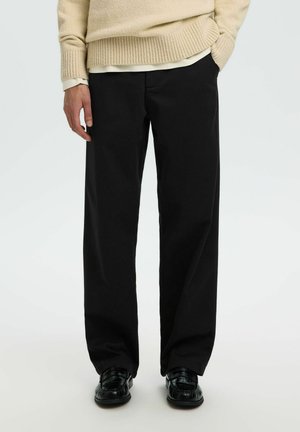 Person iført løse sorte bukser, sorte sko, beige sweater og hvid skjorte med den ene hånd i lommen.