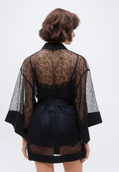 Mujer de cabello corto y castaño vista desde atrás, vestida con una bata estilo kimono de encaje negro transparente, con mangas anchas y ribete de satén.