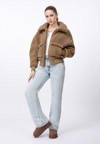 Fiatal nő barna polárdzsekiben, fehér ingben, világoskék farmerral és barna slip-on cipőben, egyszínű fehér háttér előtt áll.