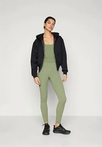 Donna in piedi che indossa un completo attivo aderente verde oliva, una felpa nera con zip e scarpe da ginnastica nere, su uno sfondo grigio uniforme.