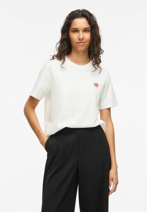Femme aux cheveux bouclés et foncés portant un t-shirt blanc avec un petit logo fraise et un pantalon noir taille haute, main dans la poche, expression neutre.
