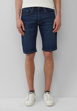 Temno modri jeans kratki hlače s ravnim krojem, petimi žepi in zavihanimi robovi, v kombinaciji z belimi supergami na nevtralni podlagi.