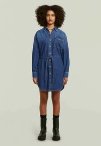 Φόρεμα πουκαμίσας από τζιν με μακριά μανίκια, γιακά, κουμπιά μπροστά, τσέπη στο στήθος και συνδυασμένη ζώνη. Το χρώμα είναι σκούρο μπλε τζιν; η υφή είναι λεία.