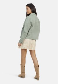 Veste en denim vert délavé avec poches latérales, associée à une robe fleurie beige et des bottes de cowboy beiges. La veste a un col classique et des poignet à bouton.