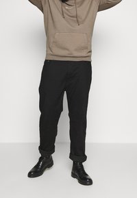 Sudadera caqui con un bolsillo frontal y cordones, combinada con pantalones negros de corte holgado remangados en los puños y botas negras de cordones.