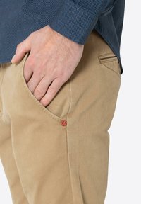 Beige Baumwollhosen mit Gesäßtasche und rotem Akzentknopf; kombiniert mit einem dunkelblauen gemusterten Hemd mit kleinen roten Punkten.