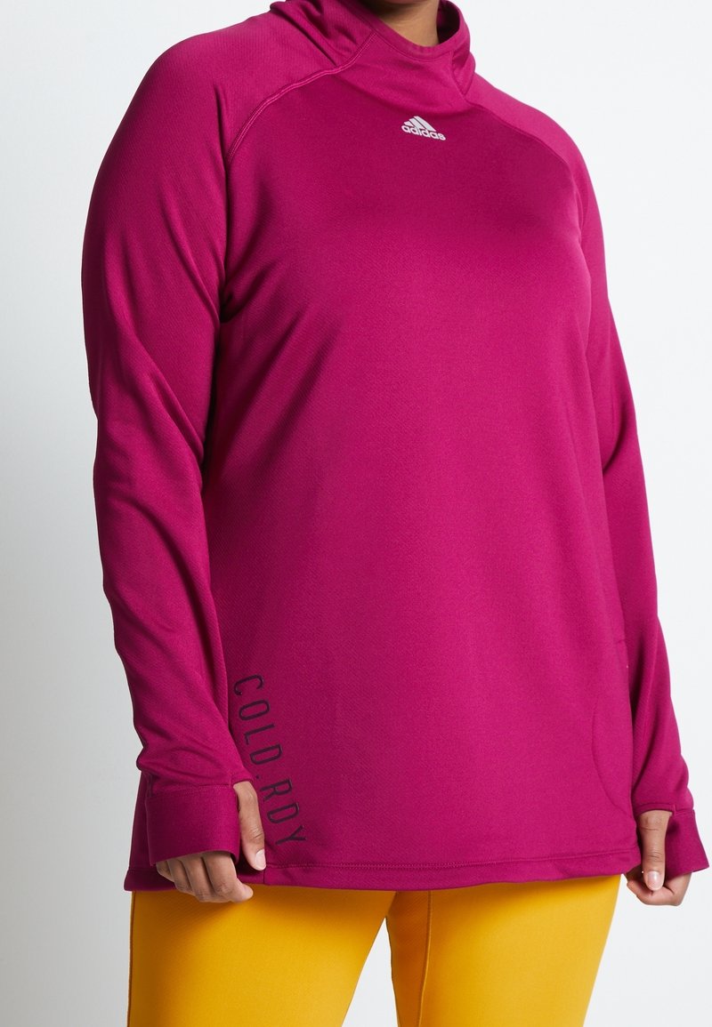 Personne portant un pull-over Adidas à manches longues magenta avec des trous pour les pouces et le texte "COLD.RDY", associé à un pantalon jaune.