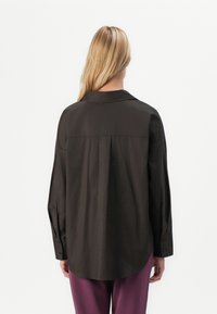 Mørkebrun skjorte med lange ermer, krage, løs passform og knapper ved håndleddene. Glatt tekstur, minimalistisk design og ingen synlige mønstre.
