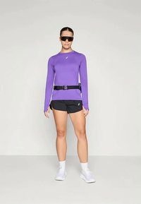 Cămașă lungă mov cu găuri pentru degete, asortată cu pantaloni scurți negri și o curea neagră. Modelul poartă ochelari de soare întunecați și pantofi albi.