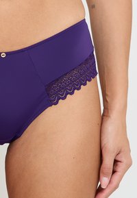 Fondodibikini viola con tessuto liscio e bordo in pizzo decorativo. Presenta un orlo scallop e una leggera lucentezza sul materiale.
