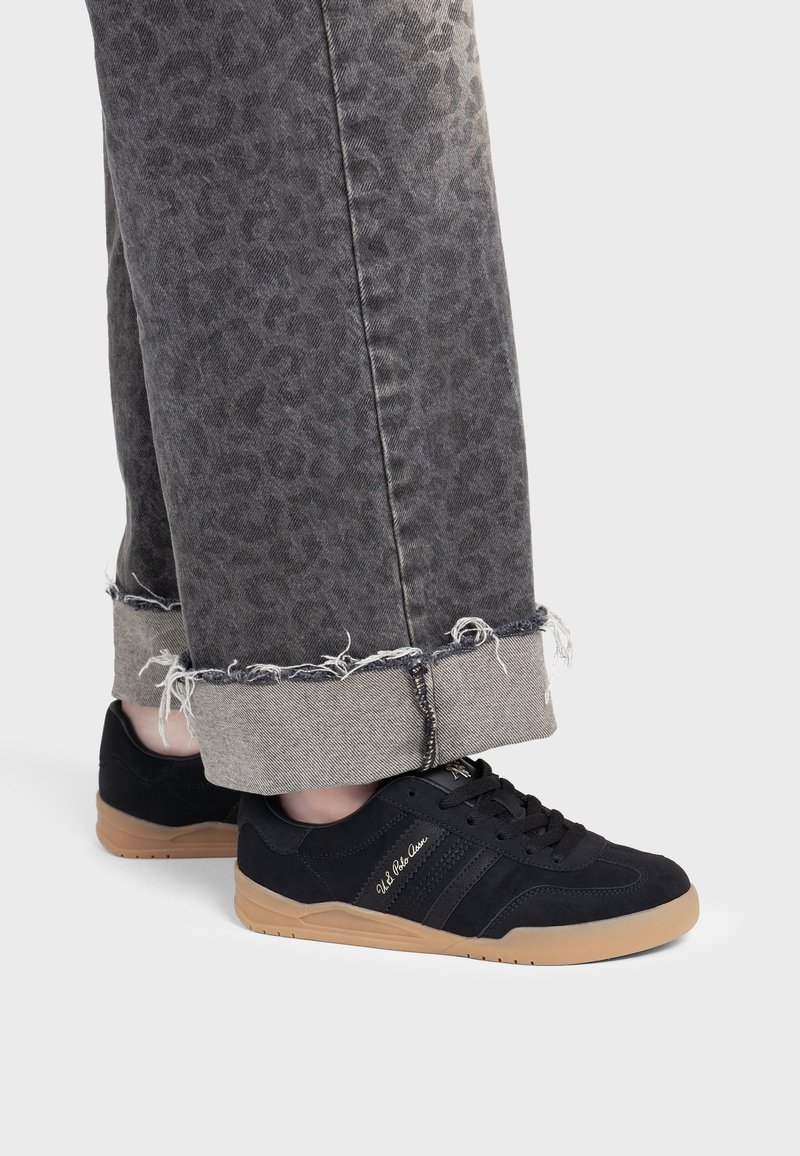 Personne portant des baskets noires avec semelles en gomme et un jean gris à imprimé léopard retroussé aux chevilles montrant des bords effilochés.