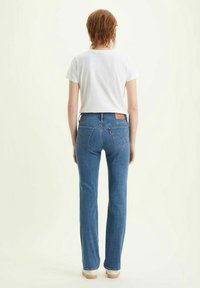 Jean denim bleu à coupe droite, cinq poches et une étiquette en cuir de marque à l'arrière de la ceinture. Porté avec un t-shirt blanc.