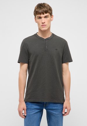 Junger Mann, der ein dunkelgraues Henley-Shirt mit kurzen Ärmeln und blaue Jeans trägt, steht vor einem schlichten hellen Hintergrund.