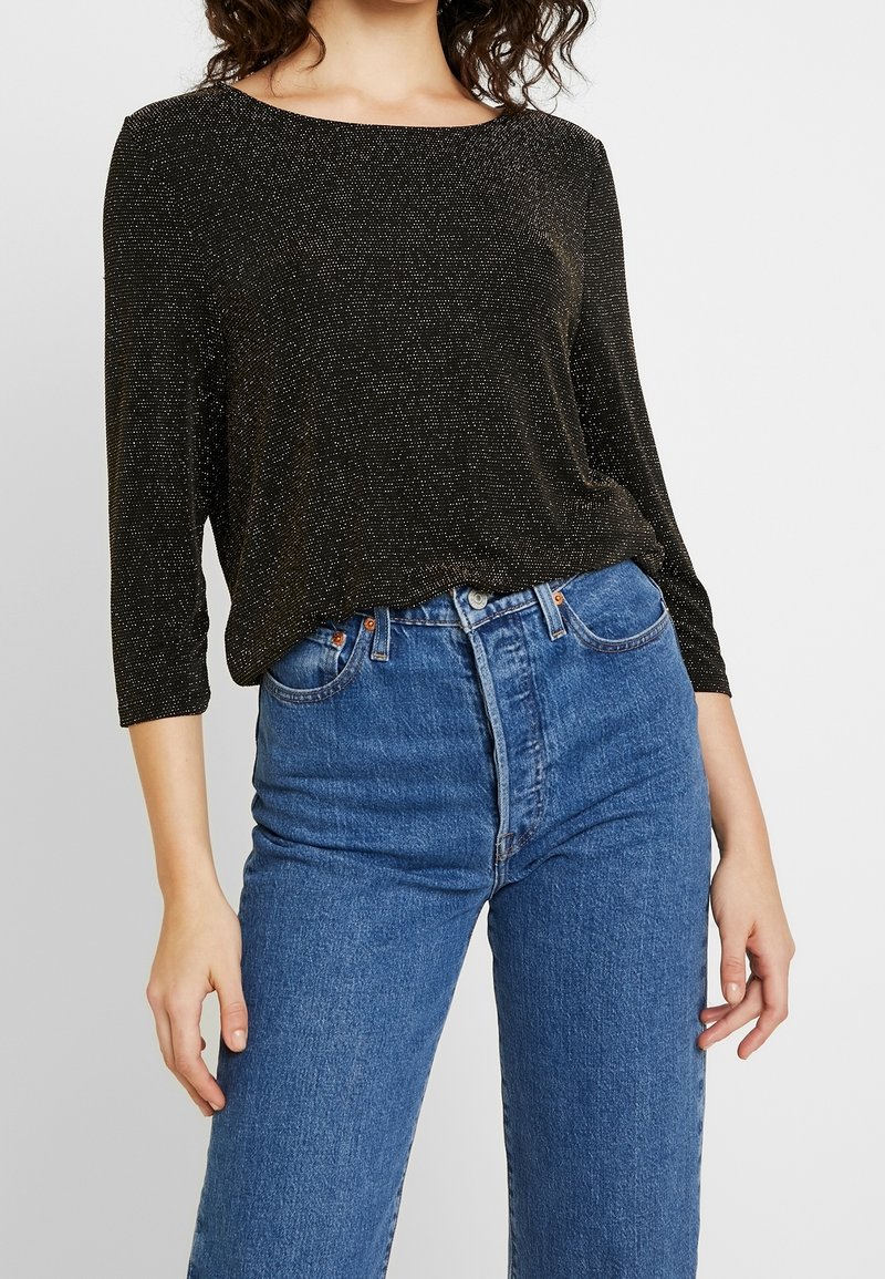 Top a maniche lunghe nero e luccicante con scollatura a barca, abbinato a jeans blu a vita alta. Il top presenta una vestibilità morbida e una texture delicata.