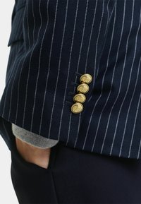 Blazer navy a righe sottili con un tessuto testurizzato, dotato di quattro bottoni dorati con design in rilievo sul polsino e un maglione grigio visibile sotto.