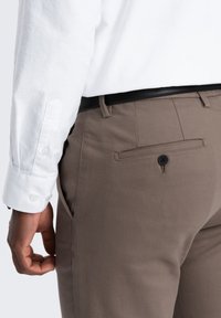 Bruine katoenen broek met een achterzak en een zwarte knop, gecombineerd met een wit shirt met knoopmanchetten.