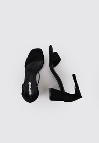 Sandalias de tacón alto de gamuza negra con diseño de jaula, punta abierta y correa en el tobillo. Presenta una plantilla texturizada y tacón cuadrado.