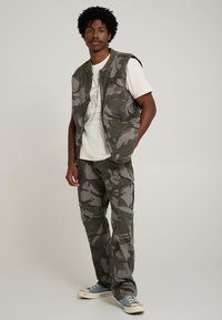 Chaleco de camuflaje y pantalones cargo a juego en tonos verdes apagados. El chaleco tiene cierre frontal con cremallera y varios bolsillos; los zapatos son de lona azul con suelas blancas.