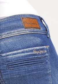 Närbild av blå Pepe Jeans jeans i denim som visar lädermärke och broderad logotyp nära bakfickan.