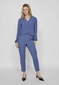 Modra bluzica z dolgimi rokavi in V-izrezom, usklajena z ozkimi hlačami. Mehka tkanina, gladka struktura, dopolnjena s črnimi čevlji s koničastimi prsti.