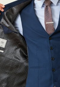 Marineblauwe gilet van textuurstof, met een patroonvoering en sluiting met vijf knopen. Versterkt met een bruine visgraatstropdas.