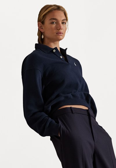 Pulover polo bleu închis cu guler, două butoane și tiv cu nervuri. Pur și simplu cu pantaloni întunecați cu talie înaltă. Detaliu subtil al logo-ului pe piept.