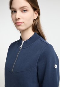 Felpa blu navy con colletto alto e chiusura con zip, caratterizzata da un design a cucitura diagonale sul davanti e un piccolo logo sulla manica.