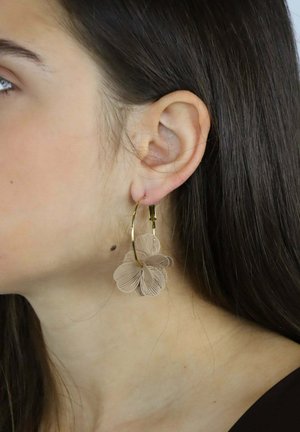 Złote kolczyki koła z dużą beżową dekoracją kwiatową, z misternie wykonanymi detalami na płatkach i gładką teksturą.