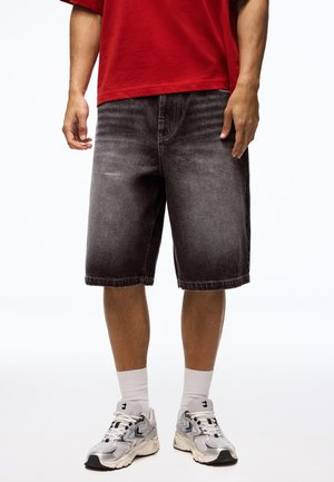 Muškarac u izblijedjelim crnim traper kratkim hlačama, bijelim sportskim čarapama, srebrnim tenisicama za trčanje i crvenoj majici na bijeloj pozadini.