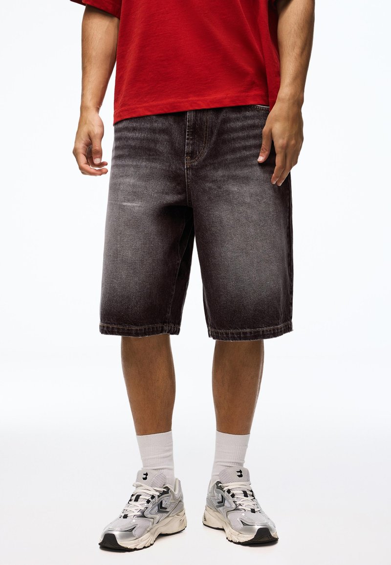 Homme portant un short en jean noir délavé, des chaussettes blanches, des chaussures de course argentées et un T-shirt rouge sur fond blanc.