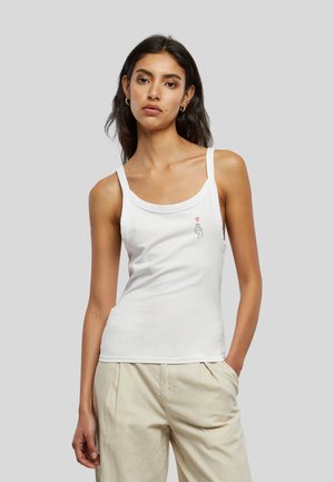 Femme aux longs cheveux foncés portant un débardeur blanc avec un petit motif main et cœur rouge et un pantalon beige sur un fond gris.