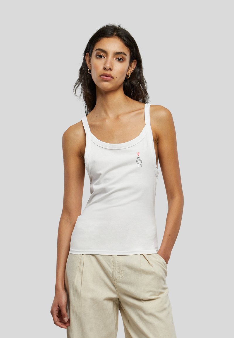 Femme aux longs cheveux foncés portant un débardeur blanc avec un petit motif main et cœur rouge et un pantalon beige sur un fond gris.