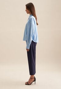 Camicia a righe blu e bianche con maniche lunghe e vestibilità comoda, abbinata a pantaloni navy su misura e sandali neri con tacco alto.