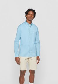 Camicia azzurra chiara con colletto e maniche lunghe, abbinata a pantaloni corti beige. Presenta un discreto logo del marchio sulla tasca del petto.