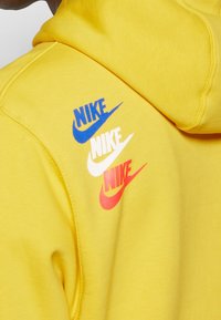 Närbild av baksidan av en gul hoodie med tre staplade Nike-logotyper i blått, vitt och rött.