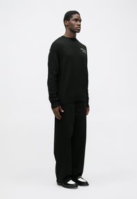 Pulover negru din tricot cu manșete și tiv ribburate, asortat cu pantaloni negri de tip slouchy. Modelul stă în fața unui fundal neutru.
