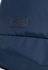 Zaino blu navy in tessuto testurizzato, con un'etichetta logo nera e un dettaglio con zip liscia. Design semplice e strutturato.