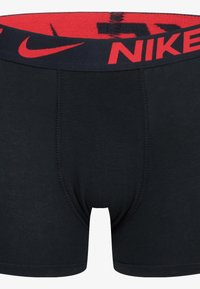Black cotton boxer briefs with a red and navy elastic waistband featuring the Nike logo prominently displayed. Smooth texture, mid-length design.