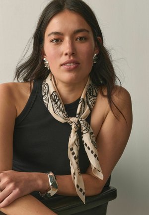 Woman with long dark hair wearing a black sleeveless top, patterned beige scarf tied at neck, silver earrings, and chunky silver bracelet sitting with folded arms.