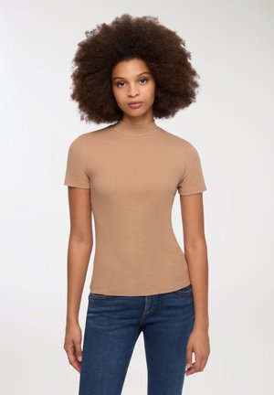 Topp a maniche corte, collo alto in tessuto beige a costine, con una forma aderente. Abbinato a jeans blu. Texture morbida, design minimal, senza motivi né dettagli.
