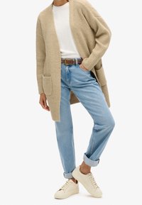 Persoon in een beige gebreide cardigan, witte śhirt, lichtblauwe opgerolde jeans, bruine riem en off-white sneakers, staand met één hand in de zak.