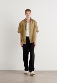 Lichtbruine short-sleeve shirt met knopen, witte tanktop eronder, zwarte broek, witte sneakers met beige zolen, tegen een effen achtergrond.