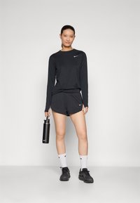 Zwarte longsleeve shirt en korte broek met een Nike-logo, gecombineerd met zwarte sportschoenen en witte sokken, terwijl er een zwarte waterfles wordt vastgehouden.